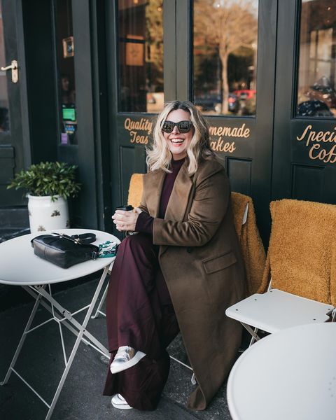 Erica Davies wearing chocolate brown coat and brown trousers sitting outside a cafe