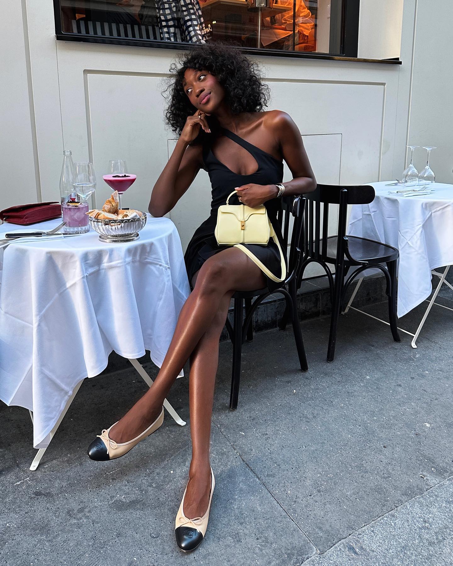 Emmanuelle Koffi siting outside at cafe table holding yellow purse wearing black dress and chanel ballet flats